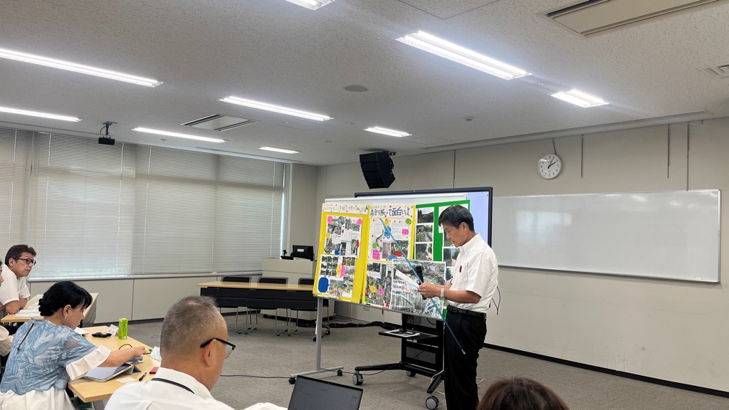 前方にスクリーンがあり、その横でマイクを持ち話す男性の話を聞いている人達を後ろから撮影した写真