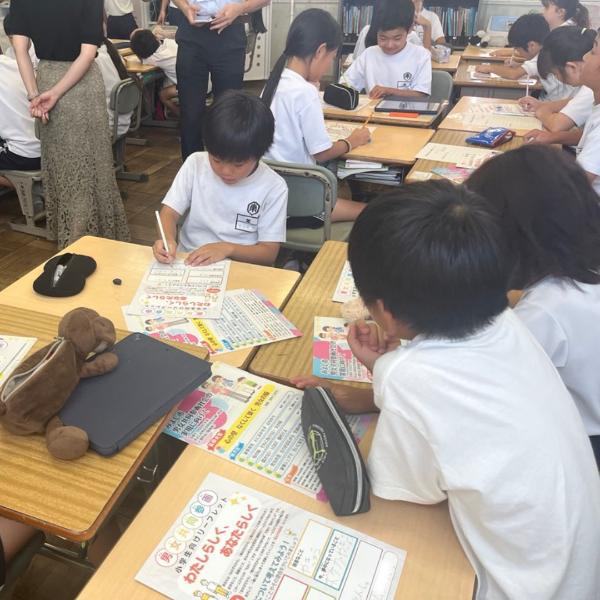 4人ずつ向き合って座っている3つのグループの児童たちが机の上に広げられたリーフレットに書き込んでいる様子の写真