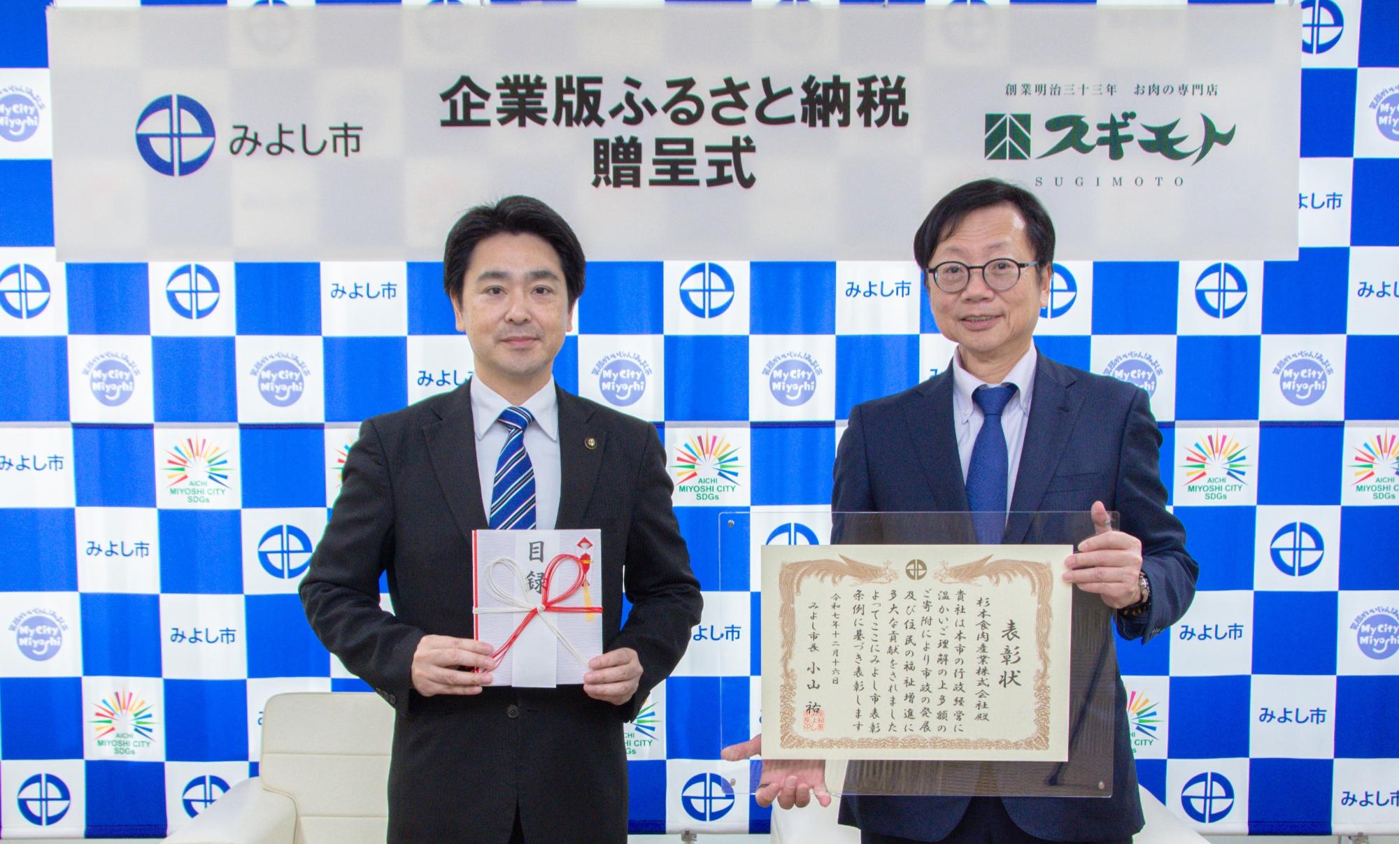みよし市のバックボードの前で市長と杉本代表取締役会長が表彰状を持ち記念撮影をしている写真