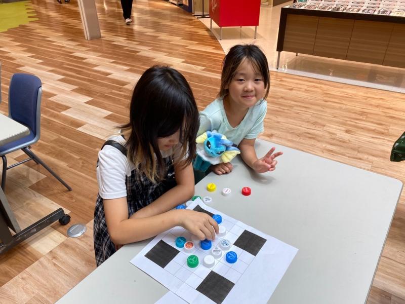 テーブルの上でペットボトルの蓋を使って遊ぶ女の子と、そのとなりでピースサインをする女の子の写真