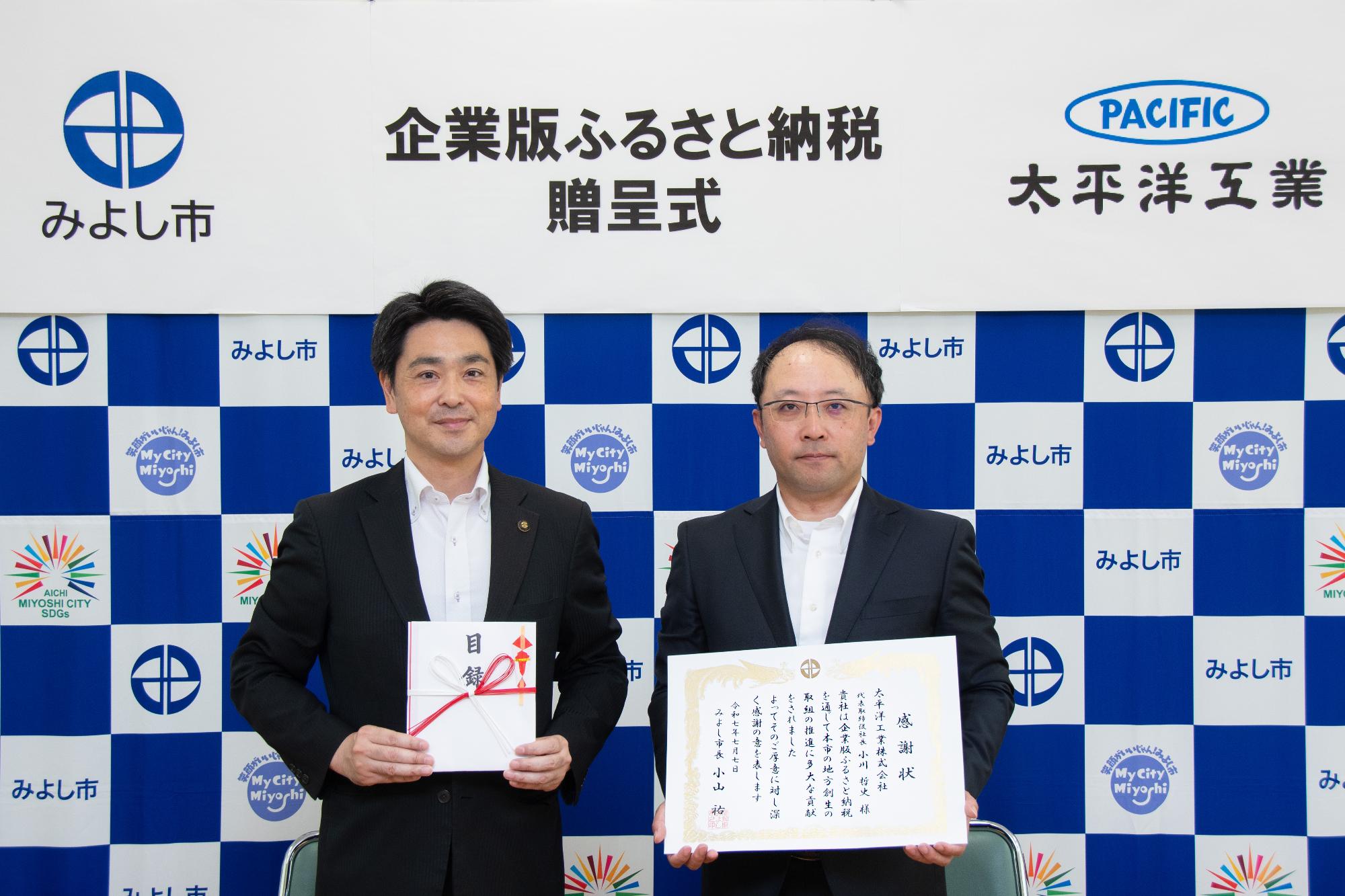 みよし市のバックボードの前で市長と小川代表取締役社長が感謝状を持ち記念撮影をしている写真