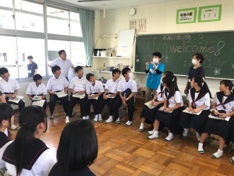 教室内で椅子を大きな円状に並べて座っている生徒たちが、水色のシャツを着て黒板の前に立っている男性の話を聞いている様子の写真