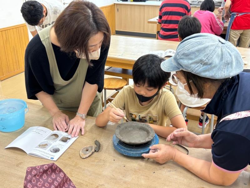 ろくろの上のお皿のふちに模様をつけている男の子と横で見ている二人の女性の写真
