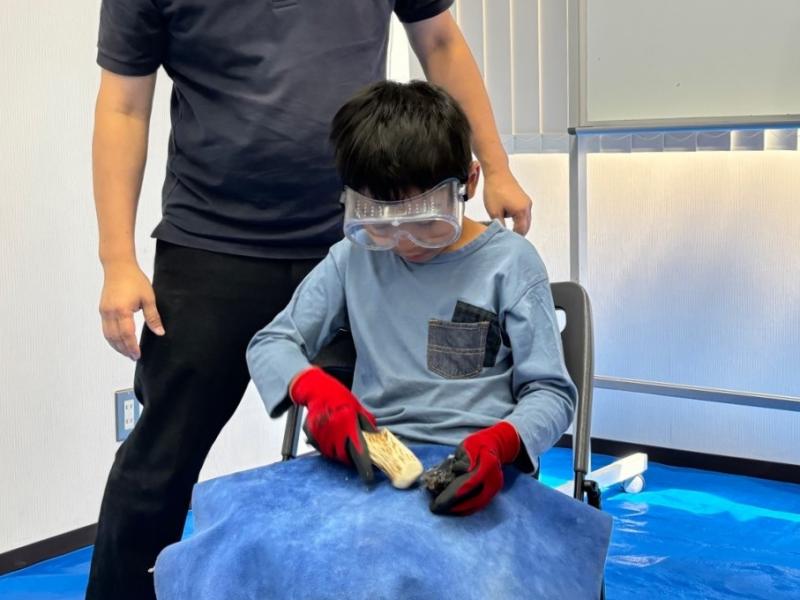 椅子に座って鹿の角を黒曜石に打ち付けている男の子と後ろから見ている男性の写真