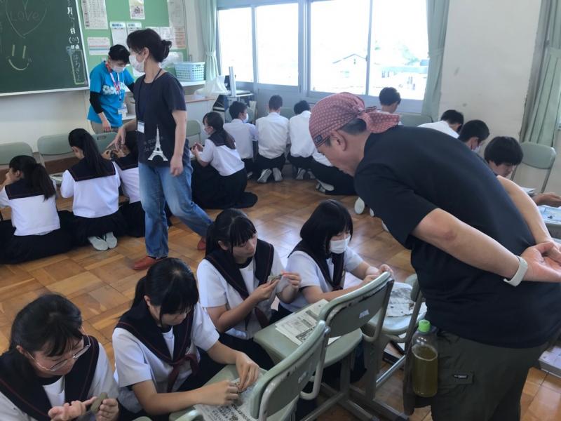 円状に並べた椅子をテーブル代わりに使って床に座って作業をしている生徒達の様子をバンダナを付けた男性が見ている写真