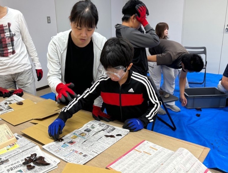 石の破片で新聞紙を切って切れ味を確かめている男の子と隣で見ている女性の写真