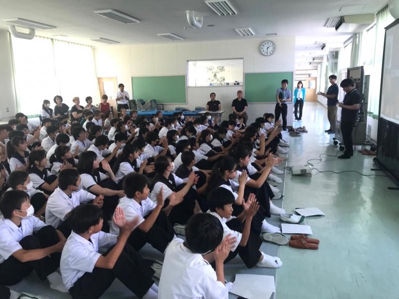 広いホールのような場所に生徒たちが並んで座り、生徒たちの前に2名の男性が立っている写真