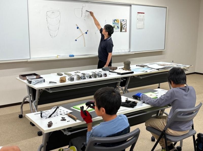 ホワイトボードに絵をかいて説明してる資料館職員男性と話を聞いている子どもたちの写真