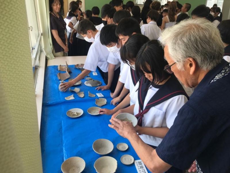 青いビニールシートが敷かれた台の上に遺跡から出土した焼き物が並べられ、中学生たちが手で触ったりしながら焼き物を見ている写真