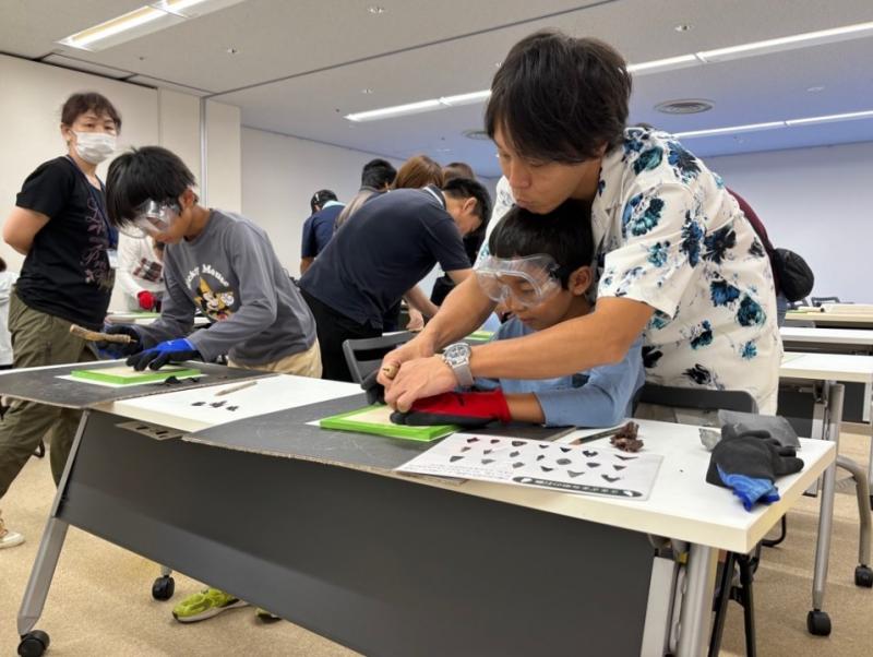 席に座っているゴーグルをつけた男の子の後ろから男性が男の子の手元を支えながら、男の子と一緒に作業している写真