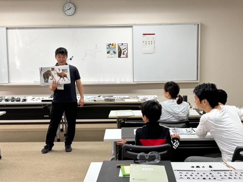 ホワイトボードの前に立って絵本を見せながら説明している資料館職員男性と話を聞いている子供たちの写真