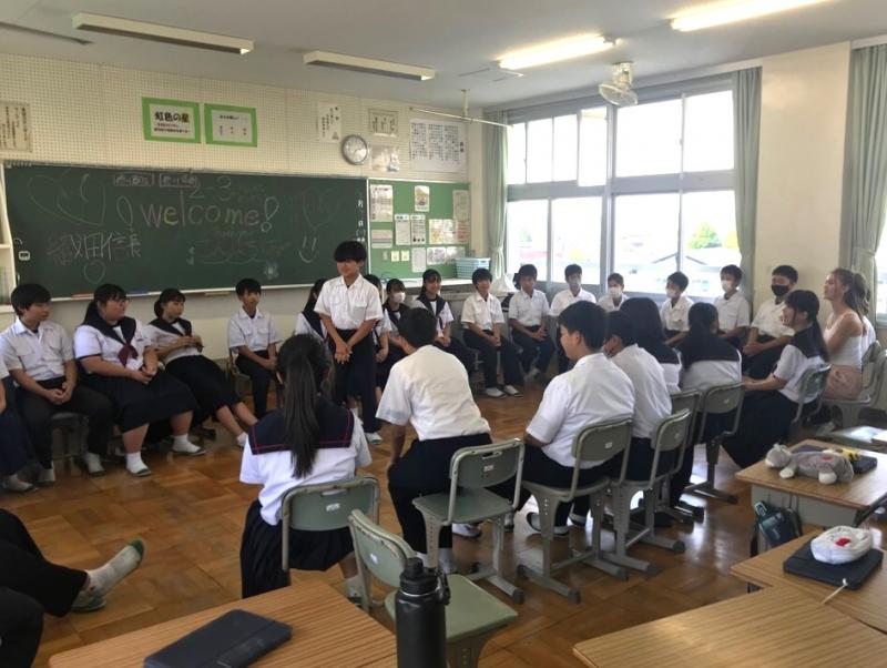黒板に「welcome」と書かれている教室内で椅子を大きな円状に並べて座っている生徒たちの中心に男子生徒が立っている写真