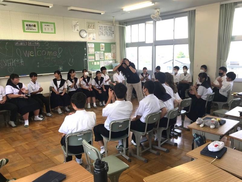 円状に椅子を並べて座っている生徒たちの中心で、コロンバス市から来た生徒が両手を頭に置いて少し前屈みになって立っている歓迎会時の写真