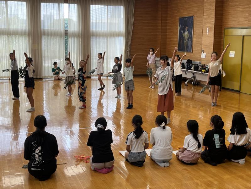左手を上にあげて整列して並んで立っている子供たちとその横で座って見ている子どもたちの写真