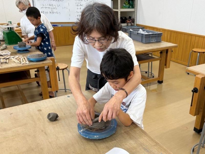 ろくろの上の焼き物の形を整えている男の子と背後から手を伸ばし、一緒に形を整えている女性の写真