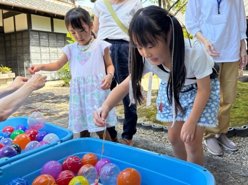 カラフルな水風船が入ったコンテナの前に立って風船釣りをしている二人の女の子の写真