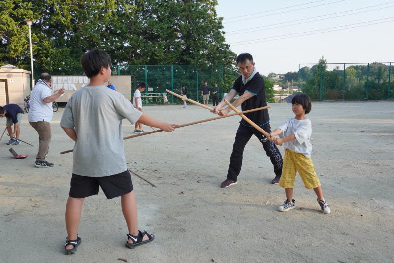 長い棒や木刀をもって打ち合いをするように向かい合って立っている子供たちと指導している男性の写真