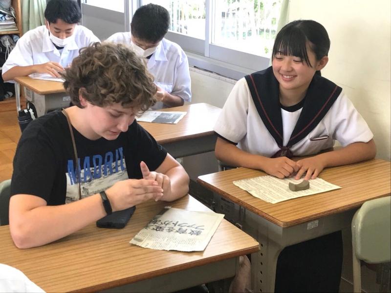 コロンバス市から来た高校生が一生懸命粘土をこねている様子を、隣の席に座っている三好中学校の女子生徒が笑顔で見守っている様子の写真