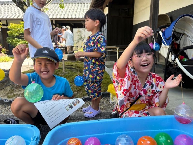 カラフルな水風船が入ったコンテナの前にしゃがんで風船釣りをしている男の子と女の子の写真