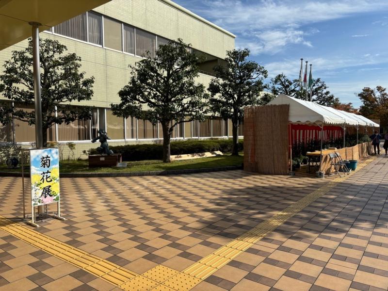 左側に「菊花展」と書かれた看板が立ち、奥にテントが並んでいる写真