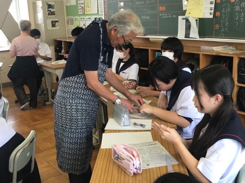エプロンを付け眼鏡をかけた年配の男性が、三好中学校の女子生徒に箸置きづくりのアドバイスをしている様子の写真