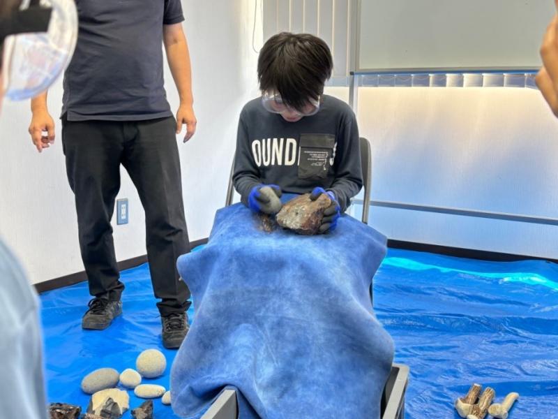 椅子に座って膝の上で黒曜石に石を打ち付けている男の子の写真