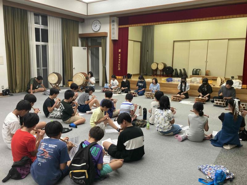 舞台の前に横一列に並んで締太鼓をたたく子供たちと向かい合って横笛を吹いている子どもたちの写真