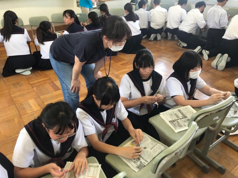 椅子をテーブル代わりに使って床に座りながら作業をしている生徒達とその様子を見て回っている女性の写真