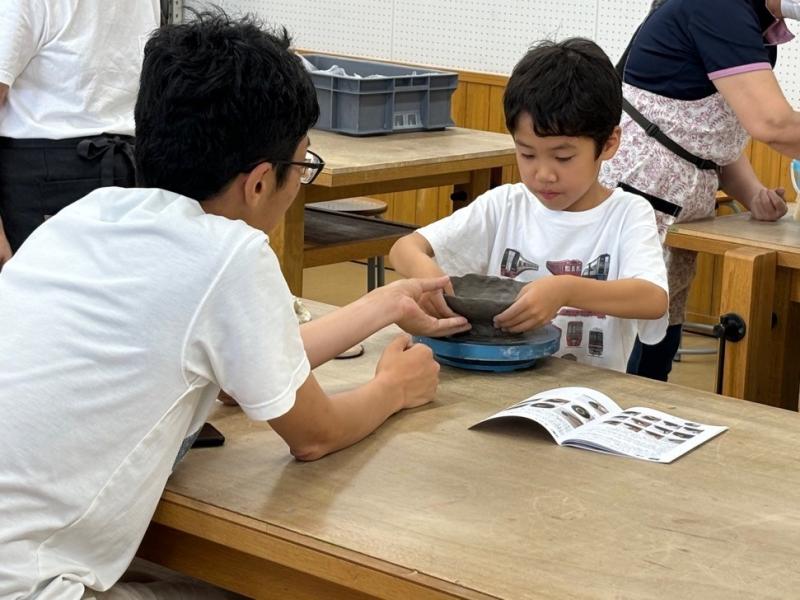 ろくろの上の焼き物の形を整えている男の子と向かい合って座っている男性の写真