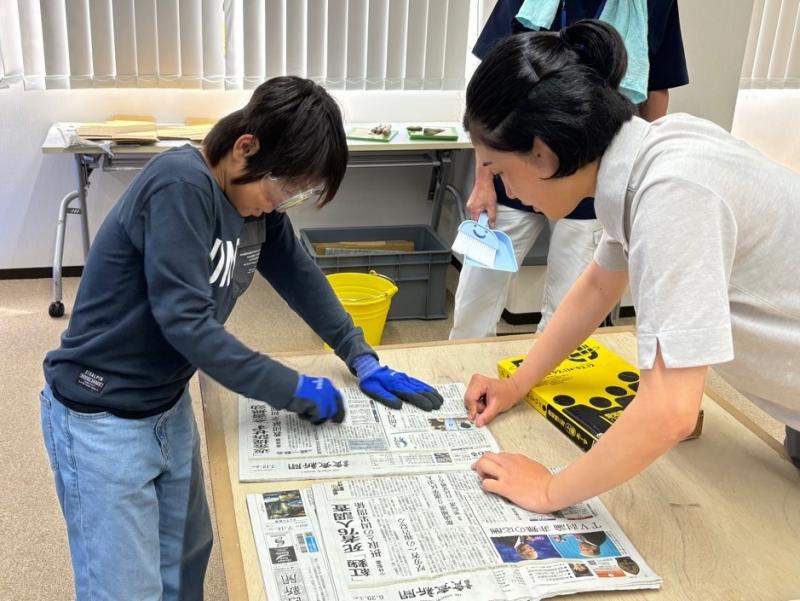 石の破片で新聞紙を切っている男の子と新聞紙を抑えている女性の写真