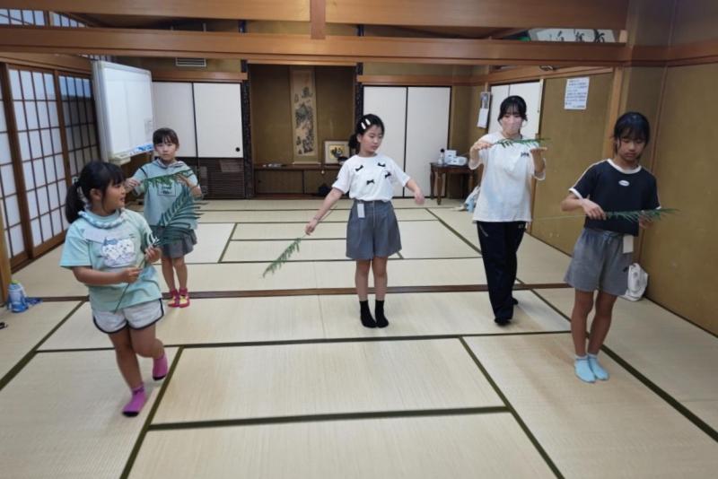 畳敷きの和室で笹の葉をもって巫女舞の練習をしている子どもたちの写真