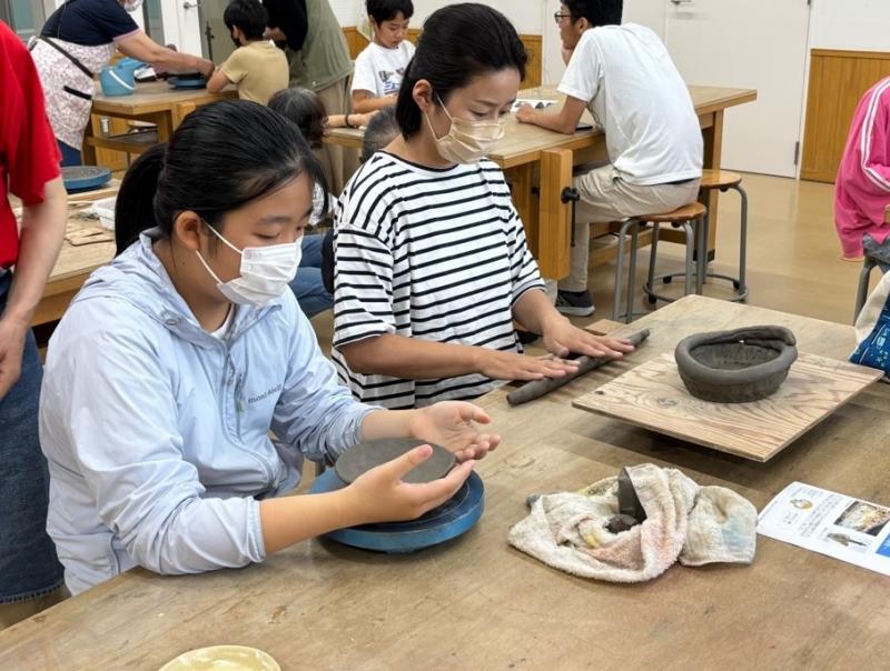 ろくろの上の焼き物の形を整えている女の子と隣で粘土を棒状に伸ばしている女性の写真