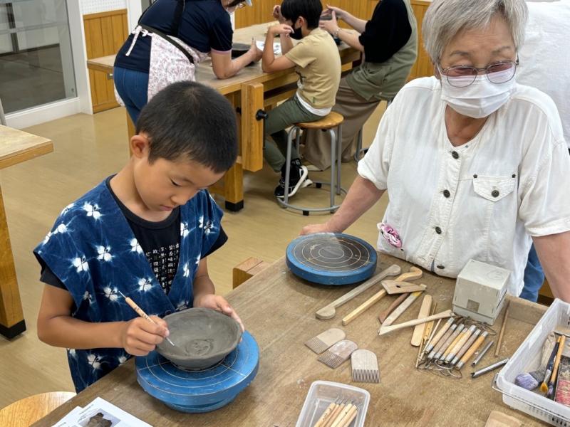 ろくろの上の焼き物に彫刻刀で模様をつけている男の子と横で見ている女性の写真