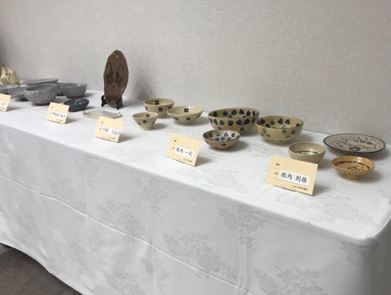 壁際に設置された白い布で覆われた長机にいろいろな種類の陶器の作品が展示されている写真