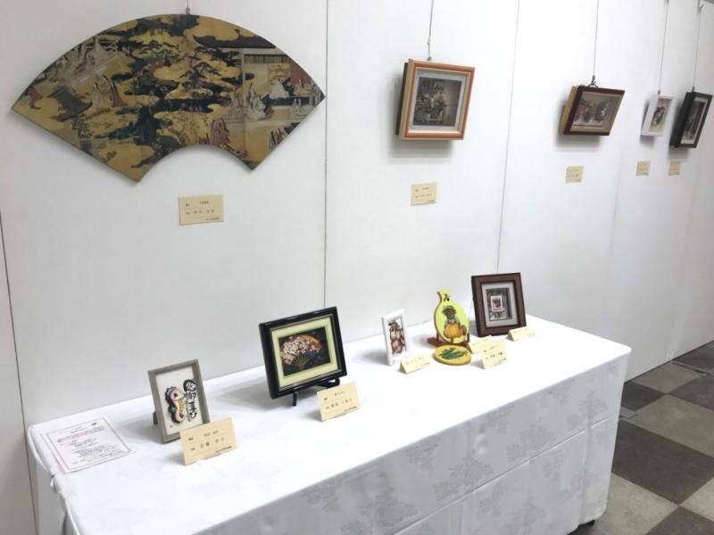 壁に扇型の作品、額に入った5つの作品、長机に5つの作品が展示されている写真