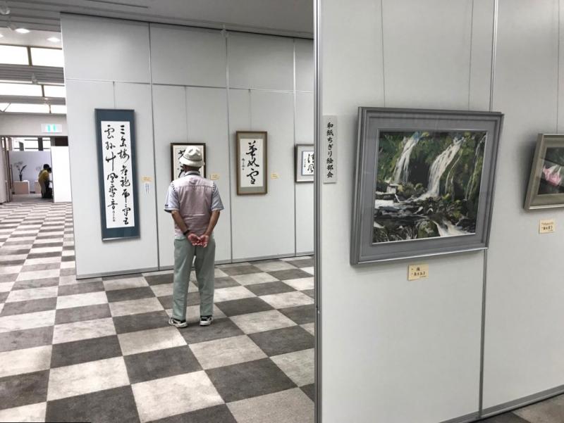 パーテーションでしきられた室内に展示されている書の前で1人の男性が立ち止まり見入っている姿を後方から写した写真