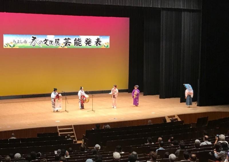 舞台に「春の文化展 芸能発表」と書かれた横断幕、マイクが2本設置された舞台の上で着物姿の5名が演技をしている写真