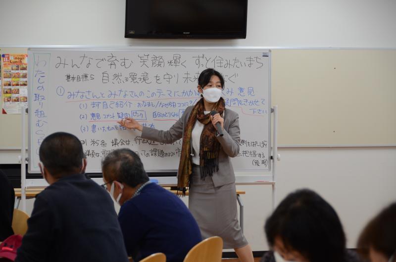 文字がたくさん書かれたホワイトボードの前に女性が立ち、マイクを持って話をしている写真