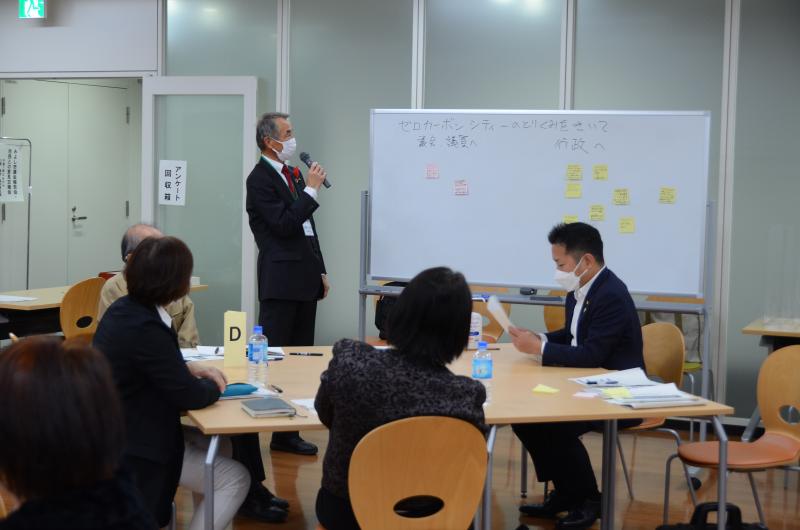 参加者達が席に着き、前方のホワイトボードの横にマイクを持って立っている男性の話を聞いている写真