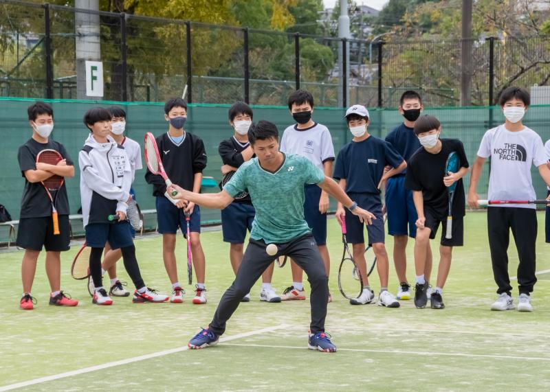 浅川さんがボールをラケットで打つ様子を横に並んだ生徒達が後ろから真剣に見ている写真