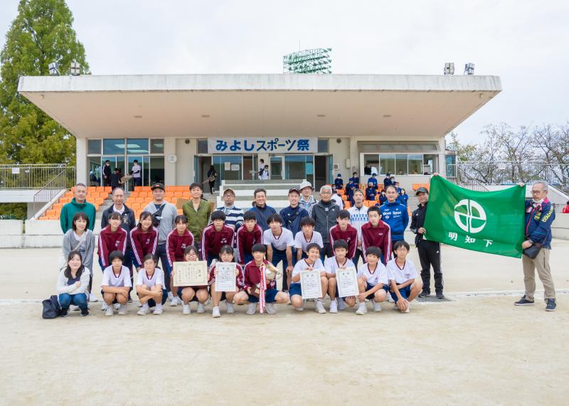 明知下の緑の旗、優勝カップ、賞状を数名が持って優勝した明知下行政区の選手達の記念写真