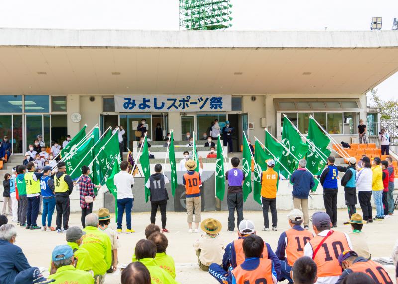「みよしスポーツ祭」と書かれ、三好町章のロゴが入った緑の旗を持った方達が並び、前に立った男性が片手を挙げ男性に向かって宣誓を行っている様子を後ろから撮影した写真