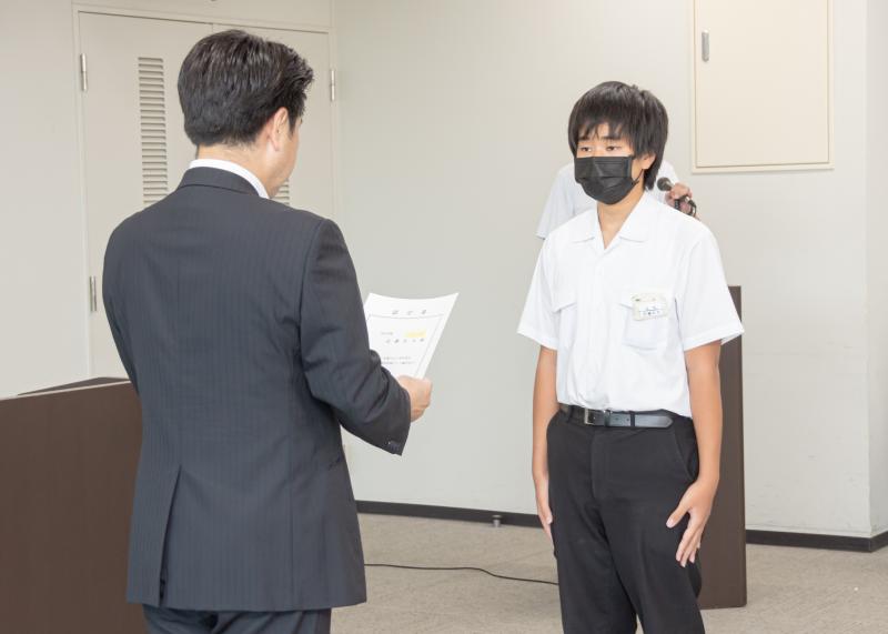 演壇前で、小山祐市長が認定書読み上げ男子中学生に手渡される様子の写真