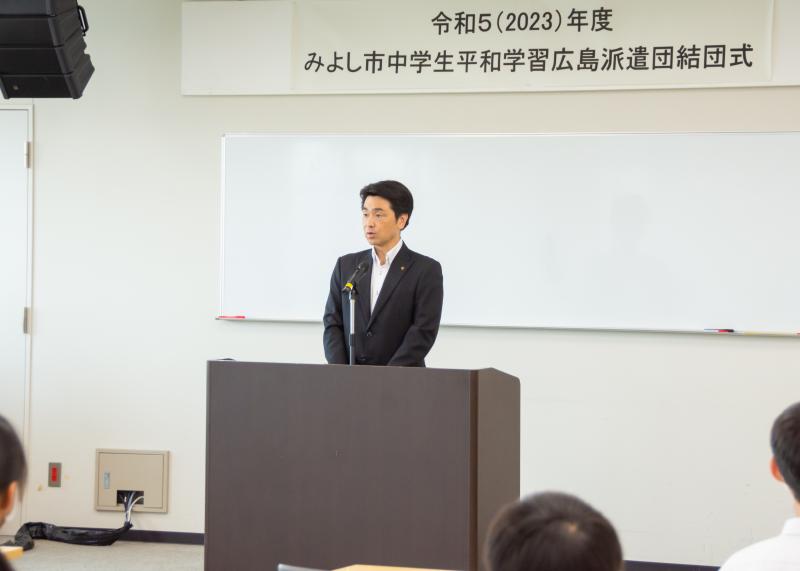 壁に「令和5(2023)年度 みよし市中学生平和学習広島派遣団結団式」と書かれた幕の前の檀上で、小山祐市長がマイクの前で話をしている様子の写真