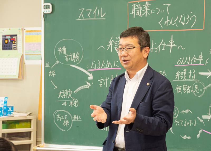 「職業についてくわしくなろう」と書かれた黒板を背に松山さんが手振りを交えながら講話を行っている写真