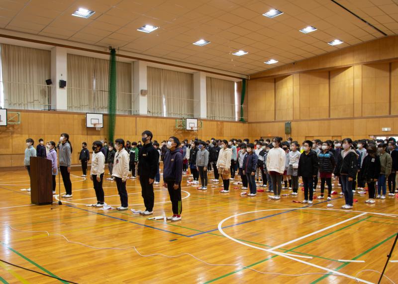 体育館に集まった三好丘小学校の児童全員が立ち、オンラインのスクリーンの方向を見ている写真
