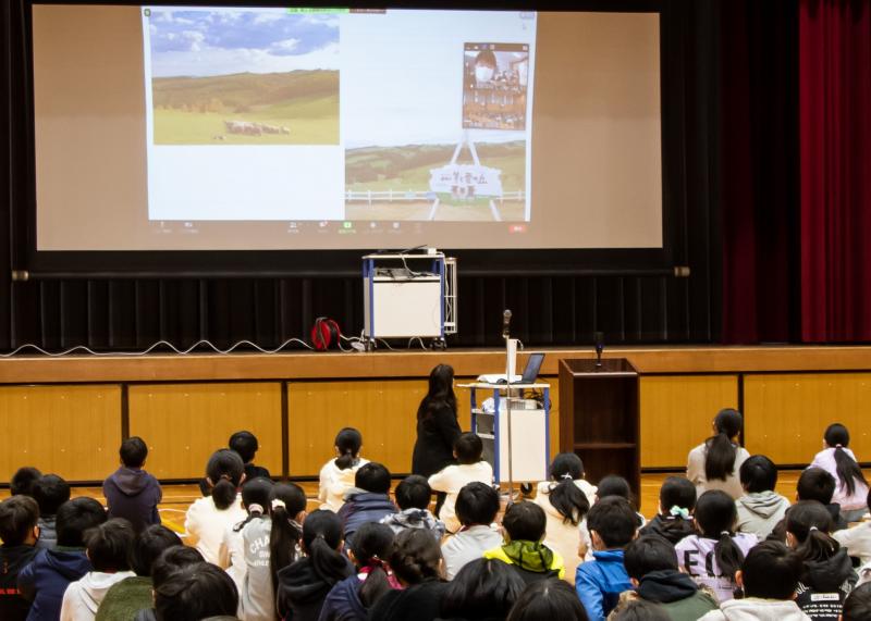 体育館のスクリーンに映された上士別小学校とのオンライン交流の画面を座って見ている児童たちの写真