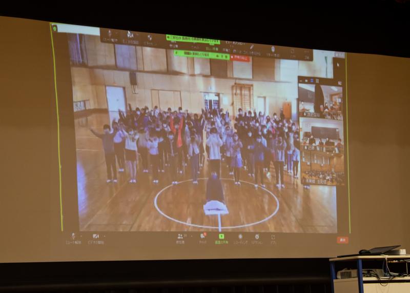 体育館に集まっている上士別小学校の児童たちがスクリーンに映し出されいる写真