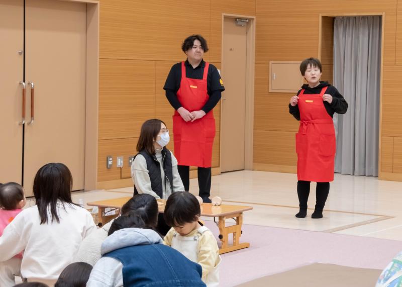 赤いエプロンを身に着けて立って話す女性と男性、その二人を座りながら見る親子の人たちの写真