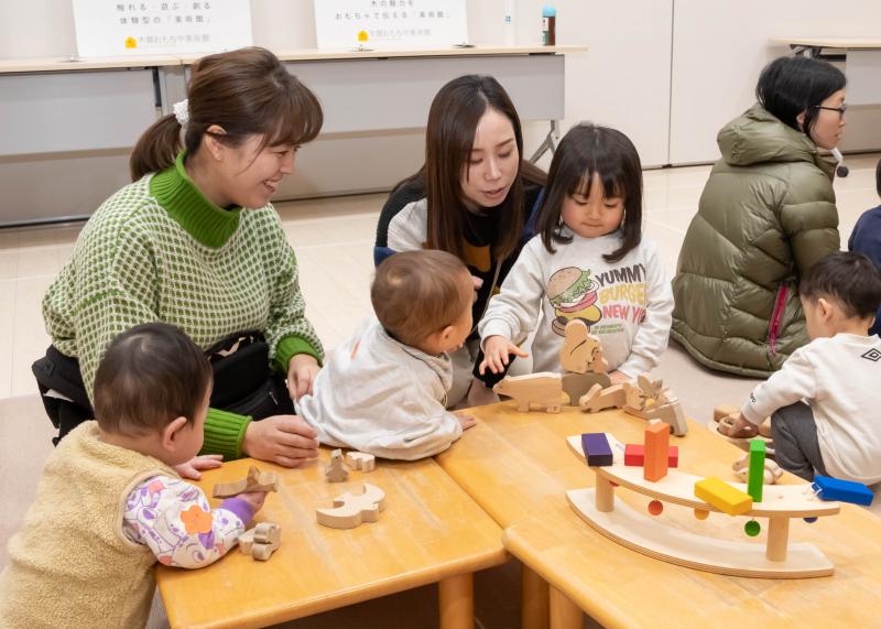 机に置かれた木製のおもちゃで遊ぶ女の子と母親を覗くようにする小さな子供とそれを見守る母親の写真